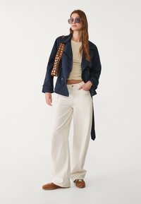 Námořnický trench coat s páskem, béžový top, krémové kalhoty širokého střihu a hnědou obuv. Oversized sluneční brýle a hnědá taška na rameno s hroty.