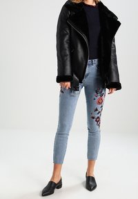 Chaqueta de motociclista de cuero negro con cuello de piel sintética, jeans azul desgastados hasta el tobillo con bordados florales coloridos a lo largo de los lados, flats negras.