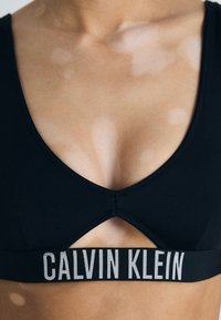Melna sporta krūšturis ar izgrieztu detaļu, kuram ir plata elastīga josla ar pelēku uzrakstu "CALVIN KLEIN", izgatavots no mīksta materiāla.