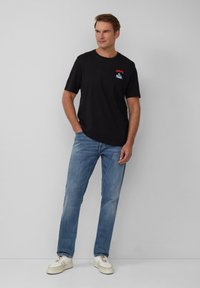 Zwarte katoenen t-shirt met rode tekst "JAMS" en een kleurrijk ontwerp op de linkerkant van de borst, gecombineerd met lichtblauwe spijkerbroek en witte sneakers.