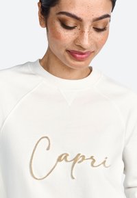 Bijela dukserica s raglan rukavima, koja na prednjoj strani ima mekani, vezeni zlatni natpis "Capri". Glatka tekstura, ležeran dizajn.