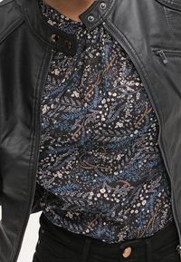 Černá kožená bunda rozepnutá, která odhaluje květinovou blůzu v námořnické modři s bílými a hnědými akcenty vyrobenou z lehkého materiálu.
