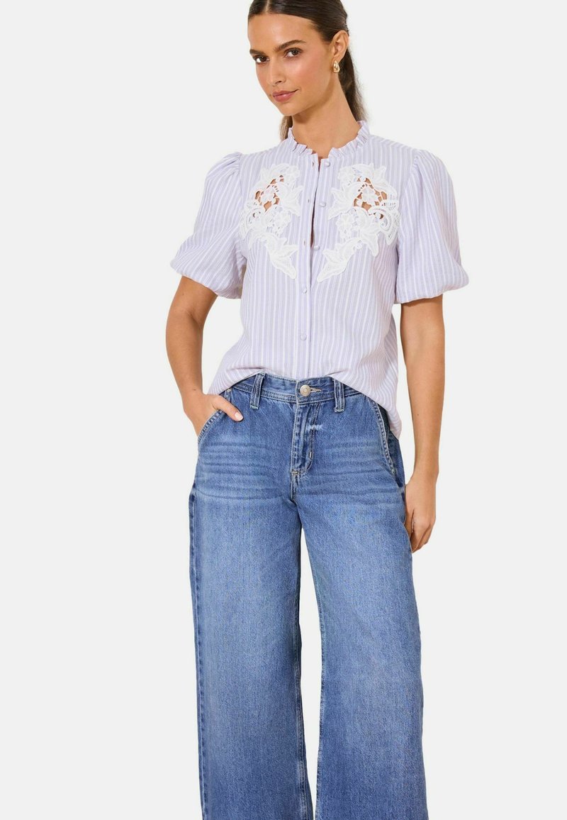 Woman wearing a lavender striped blouse with white floral lace cutouts and puff sleeves, paired with high-waisted wide-leg blue jeans.