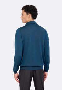 Polo a maniche lunghe di un verde scuro, con una texture morbida, colletto piatto e orlo a coste. Indossato da un modello visto di spalle.