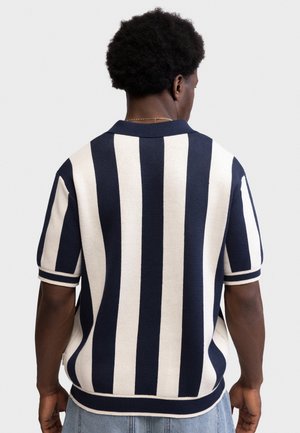 Άνδρας με κοντά κατσαρά μαλλιά που φοράει μπλε ναυτικό και λευκό πουλόβερ με κάθετες ρίγες και κοντά μανίκια, στέκεται μπροστά σε απλό φόντο.