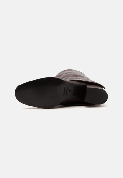 Une botte en cuir brun foncé unique, couchée sur le côté avec la semelle noire lisse et le talon bloc clairement visibles.