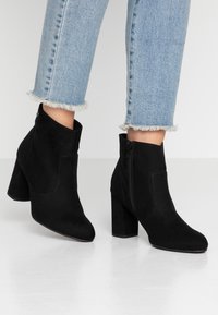 Botas de tobillo de ante negro con tacón grueso, cremallera lateral y punta afilada. Combinadas con jeans de color azul claro con dobladillos deshilachados.
