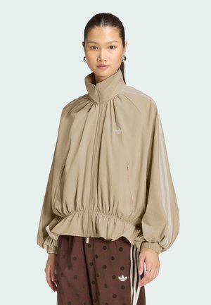 Frau trägt eine beige Zip-Jacke mit geraffter Taille und Ärmeln, kombiniert mit braunen polka-dot Hosen mit weißen Streifen und Markenlogo.