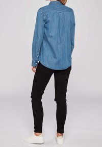 Light blue denim shirt with a button-down collar, long sleeves, a curved hem, and a relaxed fit, paired with black slim-fit pants and white sneakers.
