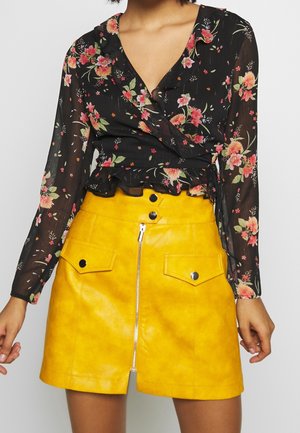 Schwarze Bluse mit floralen Mustern, langen Ärmeln und Rüschensaum, kombiniert mit einem leuchtend gelben Minirock aus Kunstleder, der über Fronttaschen und einen Reißverschluss verfügt.