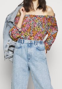 Off-shoulder top met bloemenpatroon in zwart, oranje, roze en geel. Gecombineerd met hoge lichtblauwe denim jeans. Denim jas over de schouders.