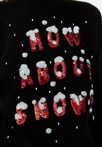 Musta villapaita, jossa on punaisilla paljeteilla kirjailtu "MITÄ SANOT LUMESTA" ja valkoisia lumihiutaleita sekä lumipipo-kuvioita koristeena.