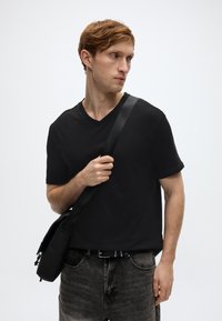 Μαύρο T-shirt με V λαιμόκοψη, φτιαγμένο από βαμβάκι, συνδυασμένο με ξεβαμμένο μαύρο τζιν σορτς. Το πρόσωπο κρατάει μια μαύρη τσάντα χιαστί. Απλό φόντο.