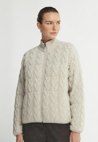 Ljusbeige kabelstickad zip-jacka med hög krage, texturerat mönster och långa ärmar. Mjukt material med avslappnad passform.