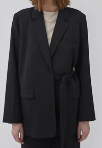 Sort blazer med bindebånd i taljen, enkelt brystlomme og to forlommer. Fremstillet af et struktureret stof med brede ærmer.