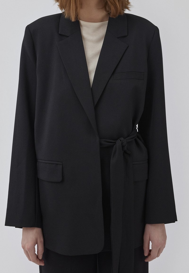 Sort blazer med bindebånd i taljen, enkelt brystlomme og to forlommer. Fremstillet af et struktureret stof med brede ærmer.
