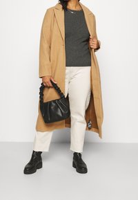 Długi płaszcz wielbłądzi, szary prążkowany sweter, kremowe spodnie, czarne masywne botki oraz czarna torebka z marszczonym uchwytem. Gładkie tekstury, dopasowany krój.