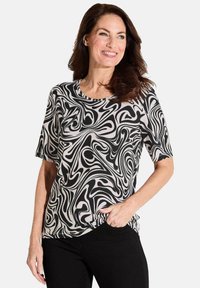 Korte mouwtop in zwart en room met een draaiend abstract patroon. Zacht materiaal, ronde halslijn en een relaxte pasvorm. Zwarte jeans zichtbaar.