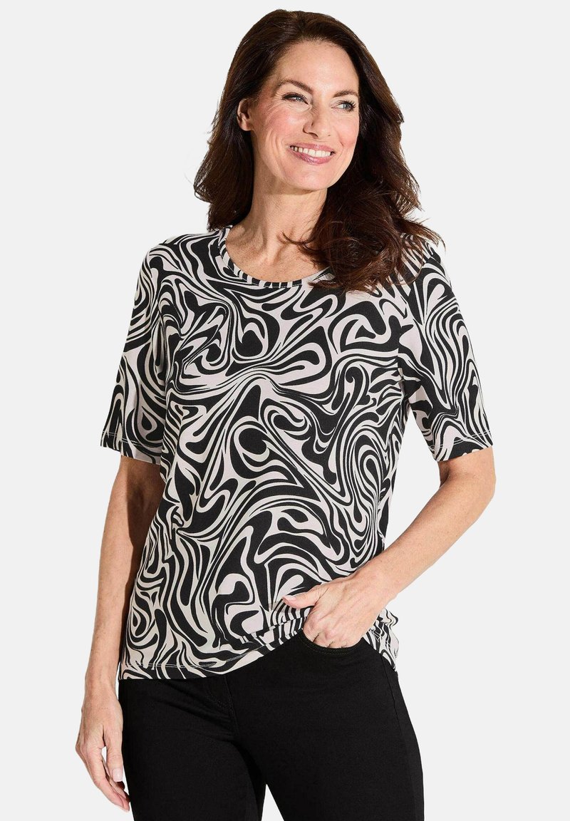 Korte mouwtop in zwart en room met een draaiend abstract patroon. Zacht materiaal, ronde halslijn en een relaxte pasvorm. Zwarte jeans zichtbaar.