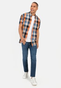 Korte mouwen geruite shirt in blauw, oranje en zwart, gecombineerd met een wit T-shirt en blauwe jeans. Afgemaakt met witte sneakers.