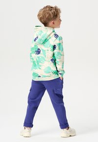 Kapuzensweatshirt mit grünen und blauen Blattmustern auf cremefarbenem Hintergrund, kombiniert mit lila Cargo-Shorts und hellen Sneakers.