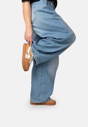 Persoon draagt wijde blauwe spijkerbroek en lichtbruine sneakers, tilt één been achterop en houdt de schoen vast met de hand.