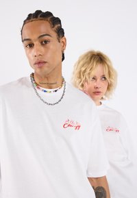 Vita bomulls t-shirts med en lös passform. Framsidan har en röd "Energy" grafik med låga-accenter. Modellen bär lager av halsband med färgglada pärlor.