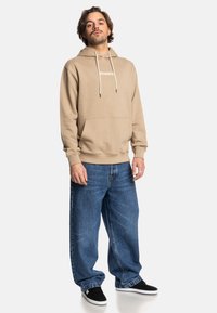 Beige hoodie met trekkoorden en een voorzak, gecombineerd met losse blauwe jeans en zwarte instappers. Model staat tegen een witte achtergrond.