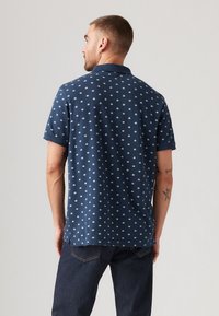 Polo shirt de marină cu modele circulare albe, mâneci scurte și guler. Materialul pare moale, cu o croială lejeră și slituri pe laterale la tiv.