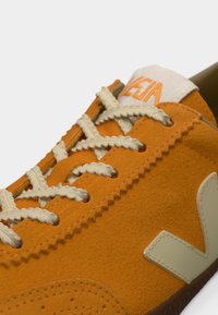 Πορτοκαλί σουέντ αθλητικό παπούτσι με μπεζ κορδόνια και διακοσμητικά στοιχεία. Διαθέτει χαρακτηριστικό υφασμάτινο σχέδιο και ελαστική σόλα. Λογότυπο στη γλώσσα.