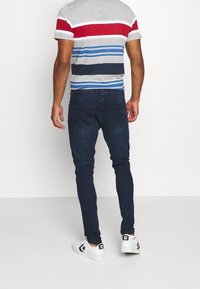 Man sedd bakifrån som bär en grå randig t-shirt med röda, blå och vita mönster, kombinerat med mörkblå jeans och vita sneakers.