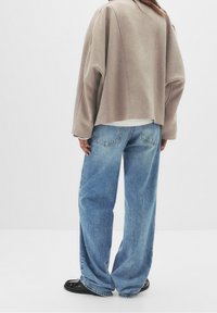 Personne portant une veste beige courte, un jean large bleu clair, des chaussures noires, debout contre un fond clair uni, de dos.