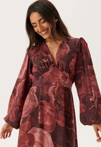 Klänning med lång ärm i mörk vinröd färg med blommönster. Har en djup V-ringning, rynkad överdel och flytande ärmar. Slät tygtextur.