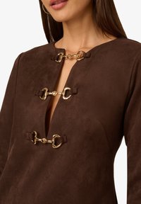 Hnědá semišová šaty s hlubokým V-výstřihem a třemi zlatými kovovými detaily na hrudníku. Hladká textura a přiléhavý střih.