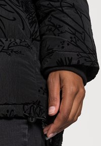 Svart pufferjacka med blommig präglad mönster. Utrustad med stickade manschetter och dragskojustering i fållen, vilket visar en texturerad yta.