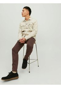 Light beige button-up jacket with two chest pockets, brown slim-fit pants, and black suede shoes with rubber soles. Seated on a stool.