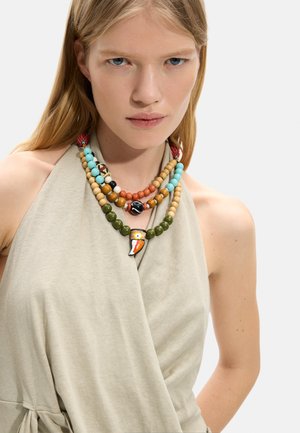 Młoda kobieta z długimi blond włosami nosi warstwowe naszyjniki z koralików, w tym jeden z zawieszką w kształcie ptaka, oraz beżowy bezrękawnik.