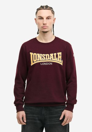 Noor mees punutud juustega, kandes burgundia punast dressipluusi kollaste ja valgete tähtedega "LONSDALE LONDON", kombineerituna tumedate teksadega.