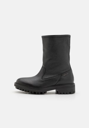 Botte mi-mollet en cuir noir avec semelle crantée épaisse, bout arrondi et logo discret Calvin Klein sur le côté du talon, présentée sur fond blanc.