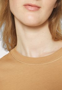 Gros plan sur le bas du visage, le cou et le haut de la poitrine d'une personne portant un t-shirt marron clair à col rond, avec une peau claire et des cheveux châtain clair visibles.