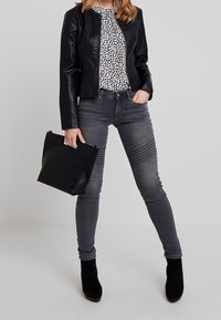 Schwarze Lederjacke über einer weißen Bluse mit schwarzen Sprenkeln, kombiniert mit grauen Jeans mit gerippten Details und schwarzen Stiefeletten. Schwarze Tasche.