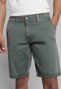 Grijs-groene katoenen shorts met opgerolde manchetten, voorzien van een bruine knoopsluiting en subtiele stikdetails, gedragen met een wit tanktop.