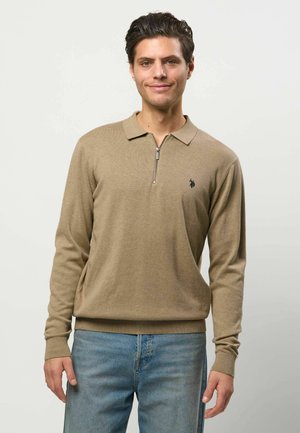 Bărbat purtând un pulover polo bej cu mâneci lungi, fermoar și un mic logo negru, asortat cu blugi albastru deschis, stând în fața unui fundal simplu.