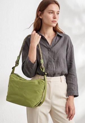 Femme portant une chemise grise et un pantalon crème tient un sac à bandoulière vert avec une sangle épaisse en corde sur un fond neutre.