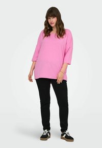 Maglietta rosa a maniche corte e vestibilità ampia, realizzata in tessuto morbido; abbinata a pantaloni neri e sneakers. Design semplice, senza motivi o accentuazioni visibili.