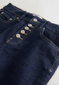 Tumši zili džinsi no denima ar pogu aizdari, kurā ir piecas metāla pogas, kontrastējošs zelta šuvums un priekšējās kabatas ar glītu dizainu.