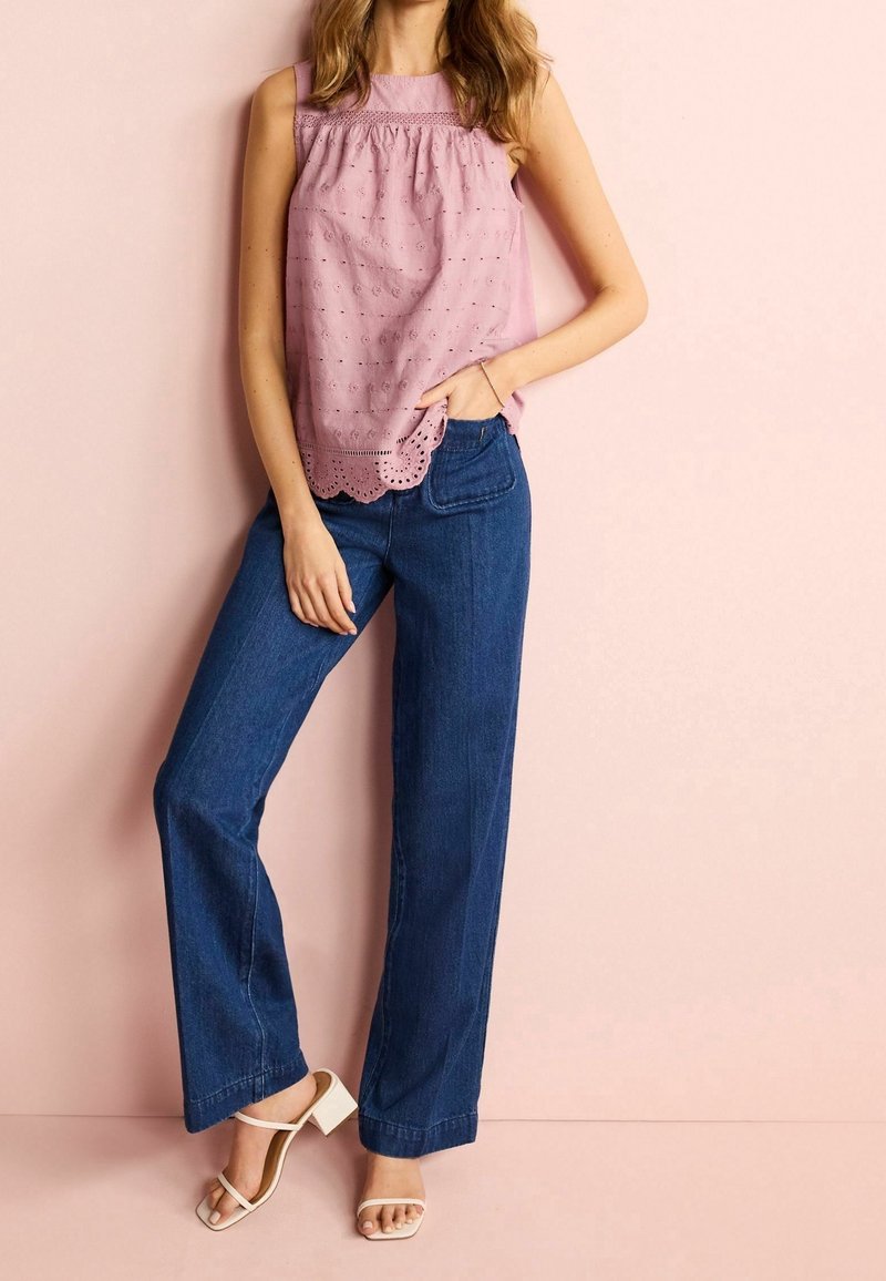 Lichtroze mouwloze top met perforaties en een geschulpte zoom, gecombineerd met middelhoge blauwe wijde jeans en witte haksandalen.