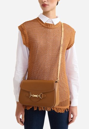 Donna che indossa una camicia bianca con bottoni e un top marrone senza maniche in maglia con orlo sfrangiato, con una borsa a tracolla in pelle marrone medio.