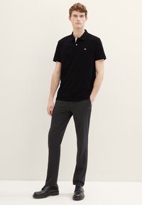 Zwarte poloshirt met korte mouwen en een klein logo, gecombineerd met grijze op maat gemaakte broek en zwarte schoenen. Eenvoudig en formeel ontwerp.