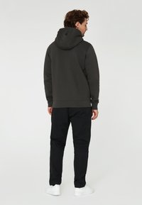 Kohlefarbener Hoodie mit einer Kordelzugkapuze, ausgestattet mit einem kleinen Logo nahe dem Hals. Kombiniert mit schwarzen Hosen und weißem Schuhwerk.